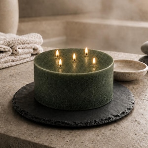 Può includere: Una grande candela rotonda verde con sei stoppini accesi è posizionata su un sottobicchiere nero strutturato. La candela è il punto focale, con un'ambientazione simile a una spa sullo sfondo, tra cui una piccola ciotola, pietre impilate e un asciugamano arrotolato.