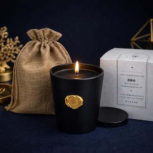 Peut inclure: Une bougie noire allumée dans un récipient noir avec un sceau doré. Une boîte blanche avec du texte et un sac en toile de jute sont à côté de la bougie. La bougie fait partie d'une collection de parfums d'intérieur.