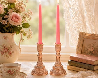 May include: Two lit pink taper candles in ornate gold candle holders. The candle holders have a detailed, embossed design. A vase of pink roses and a teacup with a floral pattern are also visible, creating a vintage aesthetic.