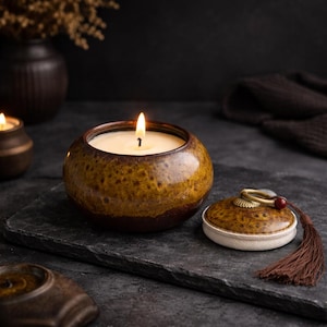 Op de afbeelding: Een brandende kaars in een ronde, bruine keramische pot met een deksel en kwast. De kaars staat op een donkergrijze ondergrond, met een donkere achtergrond en andere decoratieve items.