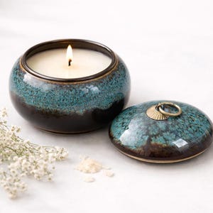 Op de afbeelding: Een brandende kaars in een teal en bruine keramische houder met een bijpassend deksel. De kaars zit in een ronde, geglazuurde keramische pot. Het deksel heeft een goudkleurige ringgreep. Op een witte ondergrond.