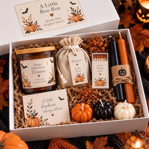 Op de afbeelding: Een witte geschenkdoos met een kaars met het label Happy Halloween Eleanor, een buideltje met een Eleanor-label, een doosje lucifers met de tekst Good Things Glow, twee dinerkaarsen en drie kleine kaarsen in de vorm van pompoenen.