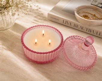 May include: A pink, three-wick candle in a decorative glass jar with a lid. The candle is lit, and the jar is partially open. The text "LIVE BEAUTIFULLY" is visible on a book in the background. A small dish with jewelry and a vase of flowers are also present.