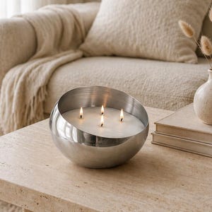 Pode incluir: Uma vela grande, prateada, em forma de tigela com quatro pavios acesos. A vela está sobre uma mesa clara e texturizada. Um sofá bege e um vaso decorativo com flores secas estão no fundo.