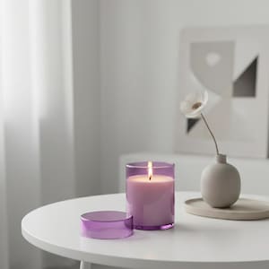 May include: A lit lavender candle in a clear glass holder with a matching lid sits on a white table. A small, white flower in a beige vase is also on the table. The candle flame is bright.