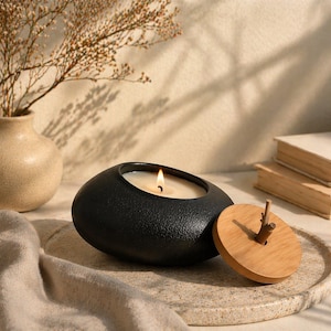 May include: A lit black candle in an oval-shaped container with a textured surface. A wooden lid with a twig handle sits beside the candle. The candle is on a speckled, light-colored tray, with a vase and books in the background.