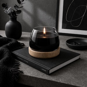 Può includere: Una candela nera accesa in un contenitore di vetro trasparente poggia su una base di legno, posta su un libro nero. Il libro è etichettato "MINIMAL LIVING" e "INTERIORS". Un vaso nero con vegetazione e un piccolo piatto nero sono sullo sfondo.