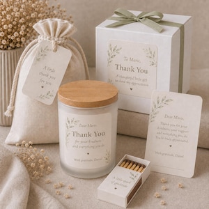May include: A gift set featuring a frosted glass candle with a wooden lid, a white gift box with a sage green ribbon, a beige drawstring pouch, a small box of matches, and two cards. All items feature botanical leaf designs and thank you text.