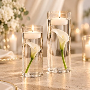 Pode incluir: Dois vasos cilíndricos de vidro transparente cheios de água, cada um contendo um lírio de calla branco submerso na água e uma vela flutuante branca acesa no topo. Os vasos têm alturas diferentes e estão posicionados lado a lado.