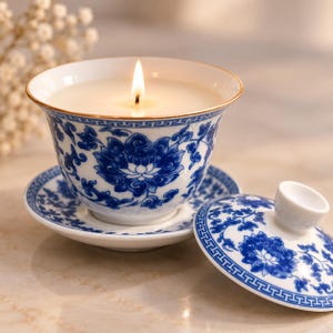Könnte beinhalten: Eine weiße Keramik-Teetassenkerze mit blauem Blumenmuster und Goldrand, die auf einer passenden Untertasse steht. Die Kerze brennt mit einer sichtbaren Flamme. Ein passender Keramikdeckel mit blauem Blumenmuster liegt daneben.