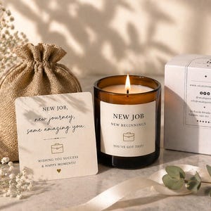 Puede incluir: Una vela encendida en un tarro de cristal marrón con una etiqueta blanca que dice "NEW JOB, NEW BEGINNINGS, YOU'VE GOT THIS!". También hay una tarjeta pequeña con el texto "NEW JOB, new journey, same amazing you" y una bolsa de arpillera. Una caja blanca está en el fondo.