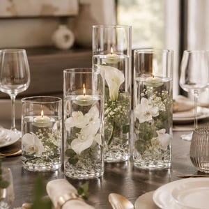 May include: Four clear cylindrical glass vases of varying heights filled with water, clear decorative beads, white orchid blossoms, green eucalyptus leaves, and baby's breath, each topped with a small, lit, round floating candle.