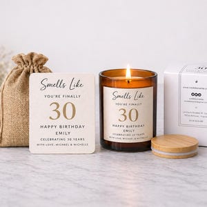 Peut inclure: Une bougie allumée dans un pot en verre brun avec un couvercle en bois, une boîte cadeau et un petit sac en toile de jute. L'étiquette de la bougie indique "Smells Like You're Finally 30 Happy Birthday Emily Celebrating 30 Years". Une carte avec le même texte est également présente.