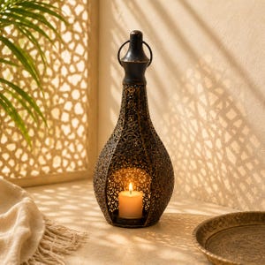 Peut inclure: Une lanterne en métal de couleur bronze foncé avec un motif floral perforé complexe et une forme en goutte d'eau. La lanterne possède une poignée supérieure et contient une bougie pilier blanche allumée projetant des ombres sur le mur.