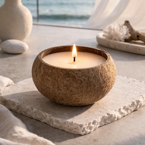 Può includere: Una candela accesa in un contenitore rotondo marrone è appoggiata su una pietra quadrata di colore chiaro. La fiamma brilla sulla cera bianca. Lo sfondo include un vaso con vegetazione e una vista sul mare, suggerendo un'atmosfera rilassante.