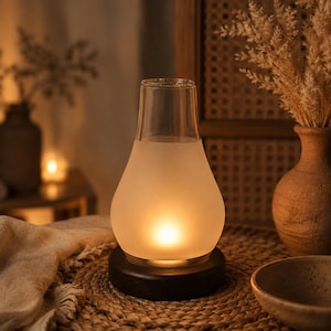 May include: A pear-shaped glass lamp featuring a clear top section and a frosted bottom section that emits a warm, glowing light. The lamp is mounted on a circular black base, creating a soft illumination effect through the translucent glass.