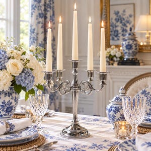 May include: A silver-toned metal candelabra featuring five arms with lit ivory taper candles. The candelabra has a central stem and curved arms with decorative scrollwork details, standing on a circular base on a table.