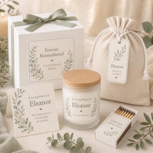 Op de afbeelding: Een herdenkingsgeschenkset met een witte doos met saliegroen lint, een beige trekkoordzakje met een gepersonaliseerd label, een matglazen kaars met houten deksel, een gevouwen kaart en een luciferdoosje, bedrukt met de naam Eleanor.