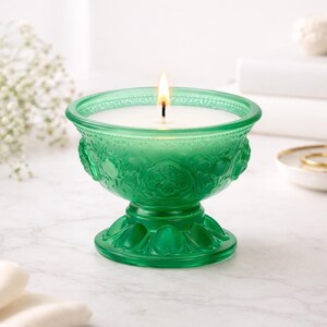 Pode incluir: Vela acesa em uma tigela de vidro verde com detalhes florais. A vela tem cera branca e um pavio. A tigela fica em uma base de pedestal. O vidro tem uma aparência vintage.