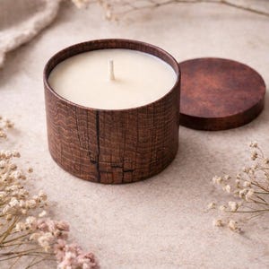 Könnte beinhalten: Eine zylindrische Kerze in einem Holzbehälter mit passendem Deckel. Die Kerze hat ein weißes Wachs und einen einzelnen Docht. Der Behälter hat ein Holzmaserungsmuster. Getrocknete Blumen sind im Hintergrund.