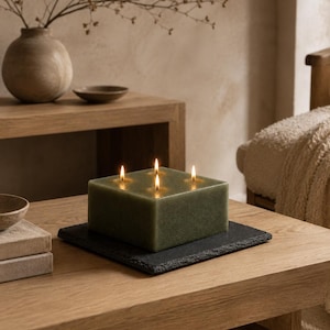 Può includere: Una candela quadrata in cera color verde salvia con quattro stoppini accesi, poggiata su una base piatta in ardesia grigio scuro con un bordo ruvido e testurizzato. La candela ha una finitura liscia e opaca ed emette una luce calda.