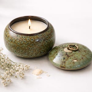 Könnte beinhalten: Eine brennende Kerze in einem runden, grünen Keramikbehälter mit Deckel. Der Behälter hat ein gesprenkeltes Muster und einen dunkelbraunen Rand. Der Deckel befindet sich neben dem Behälter und hat einen goldfarbenen Ringgriff.