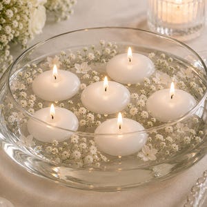 Op de afbeelding: Een heldere glazen kom gevuld met water, witte drijfkaarsen en kleine witte bloemen. Zes brandende kaarsen creëren een zachte, warme gloed. De opstelling staat op een wit tafelkleed, geschikt als decoratief middelpunt.