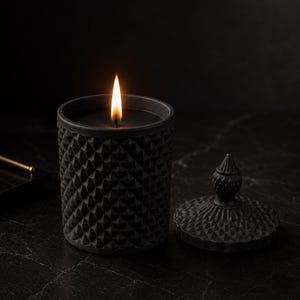 Peut inclure: Une bougie noire dans un récipient texturé à motif géométrique avec une mèche allumée, accompagnée d'un couvercle amovible assorti doté d'un fleuron pointu. La cire est noire et l'extérieur présente un motif en relief en losanges.