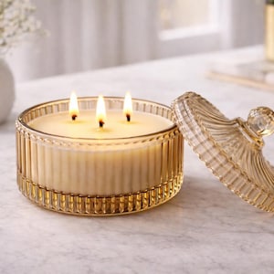 May include: A lit, three-wick candle in a clear, amber-colored glass container with a matching lid. The candle sits on a marble surface, with a vase of white flowers in the background. The candle's flame is bright.