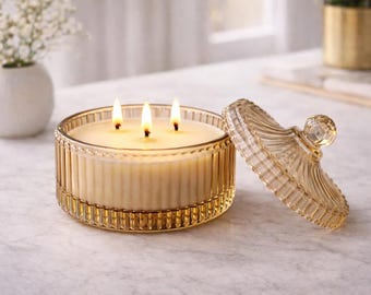 May include: A three-wick candle in a round, amber-tinted glass jar with vertical ribbing. The candle features a matching glass lid with a spherical knob, placed beside the jar. The wax is off-white, and the three wicks are lit with small flames.