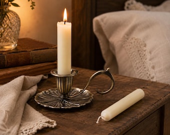 Puede incluir: Un candelabro metálico con base estriada y asa curva, que sostiene una vela cónica de color blanco roto encendida. Una segunda vela cónica de color blanco roto, sin encender, yace horizontalmente sobre la superficie junto al candelabro de acabado oscuro.