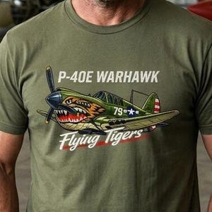 May include: An olive green short-sleeved t-shirt featuring a graphic print of a P-40E Warhawk aircraft with the text P-40E WARHAWK and Flying Tigers printed in white and red lettering across the chest of the garment.