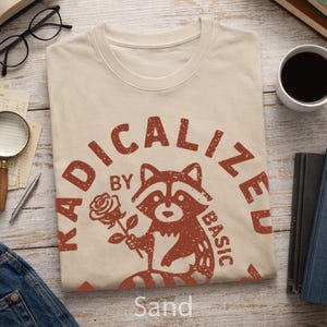 Puede incluir: Una camiseta color arena con un gráfico desgastado de color óxido de un mapache sosteniendo una rosa. El texto RADICALIZED BY BASIC está impreso en un arco circular alrededor del mapache. El diseño tiene una apariencia texturizada y desgastada.