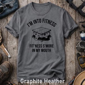 Può includere: T-shirt grigio grafite melange con la scritta "I'M INTO FITNESS" sopra una grafica di un s'more. Sotto la grafica, la scritta "FIT'NESS S'MORE IN MY MOUTH". La maglietta è in materiale morbido.