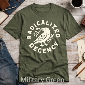 Puede incluir: Una camiseta de manga corta color verde militar con un gráfico circular en color blanco roto. El diseño incluye una ilustración central de un pájaro sosteniendo una pequeña rama, rodeado por el texto RADICALIZED BY BASIC DECENCY en negrita.