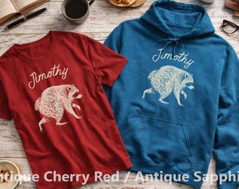 May include: A dark red short-sleeve t-shirt and a blue long-sleeve hooded sweatshirt. Both garments feature a white graphic print of a raccoon and the text Jimothy written in a cursive script above the animal illustration on the front chest.