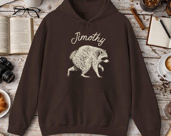 Peut inclure: Un sweat-shirt à capuche marron foncé avec un motif crème représentant un raton laveur en marche. Au-dessus du raton laveur, le nom Jimothy est imprimé dans une police cursive. Le vêtement comprend une poche kangourou et des manches longues.