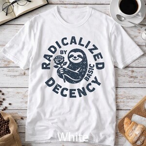 May include: A white short-sleeved t-shirt featuring a dark blue distressed graphic of a sloth holding a rose. The circular text surrounding the sloth reads "RADICALIZED BY BASIC DECENCY" in a bold, sans-serif font with a weathered texture.