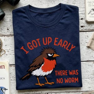 May include: A navy blue t-shirt featuring a graphic of a grumpy-looking brown and orange bird with the text "I GOT UP EARLY" arched above the bird and "THERE WAS NO WORM" printed below the bird in a bold, distressed orange font.