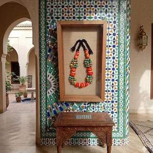May include: A framed necklace with a distinctive design. The necklace is composed of colourful beads in orange, green, and yellow. The frame is wooden, and the backdrop is a neutral linen. The necklace is displayed against a wall with detailed tilework.