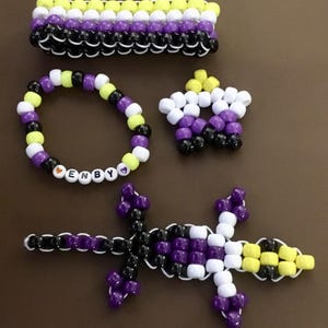 May include: A collection of handmade beaded accessories in non-binary pride colors: yellow, white, purple, and black. Includes a bracelet with "ENBY" spelled out, a star, a bracelet, and a lizard-shaped figure.