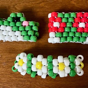 May include: Three handmade beaded bracelets. One is green and white, shaped like a frog. Another is a watermelon design with red, green, and white beads. The third bracelet is white, green, and yellow.