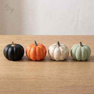 May include: A set of four decorative pumpkins arranged in a row. The first is matte black, the second is bright orange with subtle speckles, the third is a distressed off-white, and the fourth is a soft sage green. Each has a metallic gold stem.