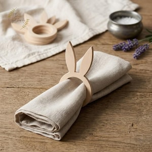 May include: A beige linen napkin held by a wooden bunny ear napkin ring. Another ring and napkin are in the background. The items are on a wooden table. A small metal bowl of salt and lavender sprigs are also visible.