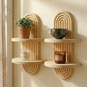 May include: Two wooden wall shelves with an arched design. Each shelf holds decorative items: a potted plant, a ceramic bowl, and two candles in glass jars. The shelves are mounted on a light-colored wall, creating a minimalist aesthetic.