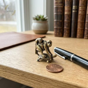 Op de afbeelding: Een kleine, messingkleurige apenfiguur die een baby-aap op zijn rug draagt, bovenop een schildpad. Het beeldje staat op een houten oppervlak, naast een cent en een zwarte pen. De achtergrond omvat een raam, boeken en een kleine potplant.
