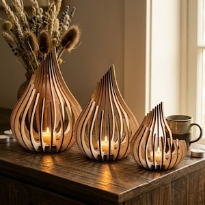 Op de afbeelding: Drie houten kandelaars met een druppelontwerp. De houders zijn gemaakt van lichtgekleurd hout en hebben verticale lamellen. Elke houder bevat een brandende kaars. De houders staan op een houten oppervlak.