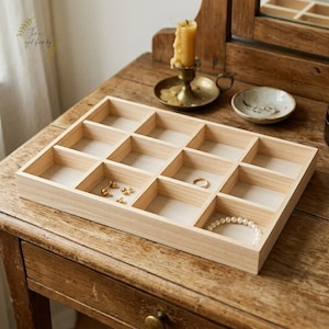 May include: A light-colored wooden jewelry tray with multiple compartments. The tray holds various jewelry items, including a gold ring, earrings, and a pearl bracelet. The tray sits on a wooden table with a candle and small dish.