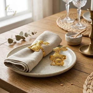 Op de afbeelding: Twee goudkleurige servetringen in de vorm van een konijn op een wit gespikkelde plaat met een beige linnen servet. De ringen hebben de vorm van een konijn. De plaat staat op een houten tafel.