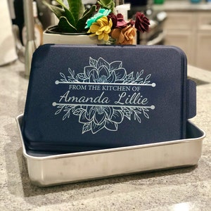 An aluminum cake pan with a navy blue lid sits on a grey marble kitchen counter, showing that the navy lid is laser engraved with a Magnolia design and the words From the Kitchen of and a name, indicating that these are personalized.